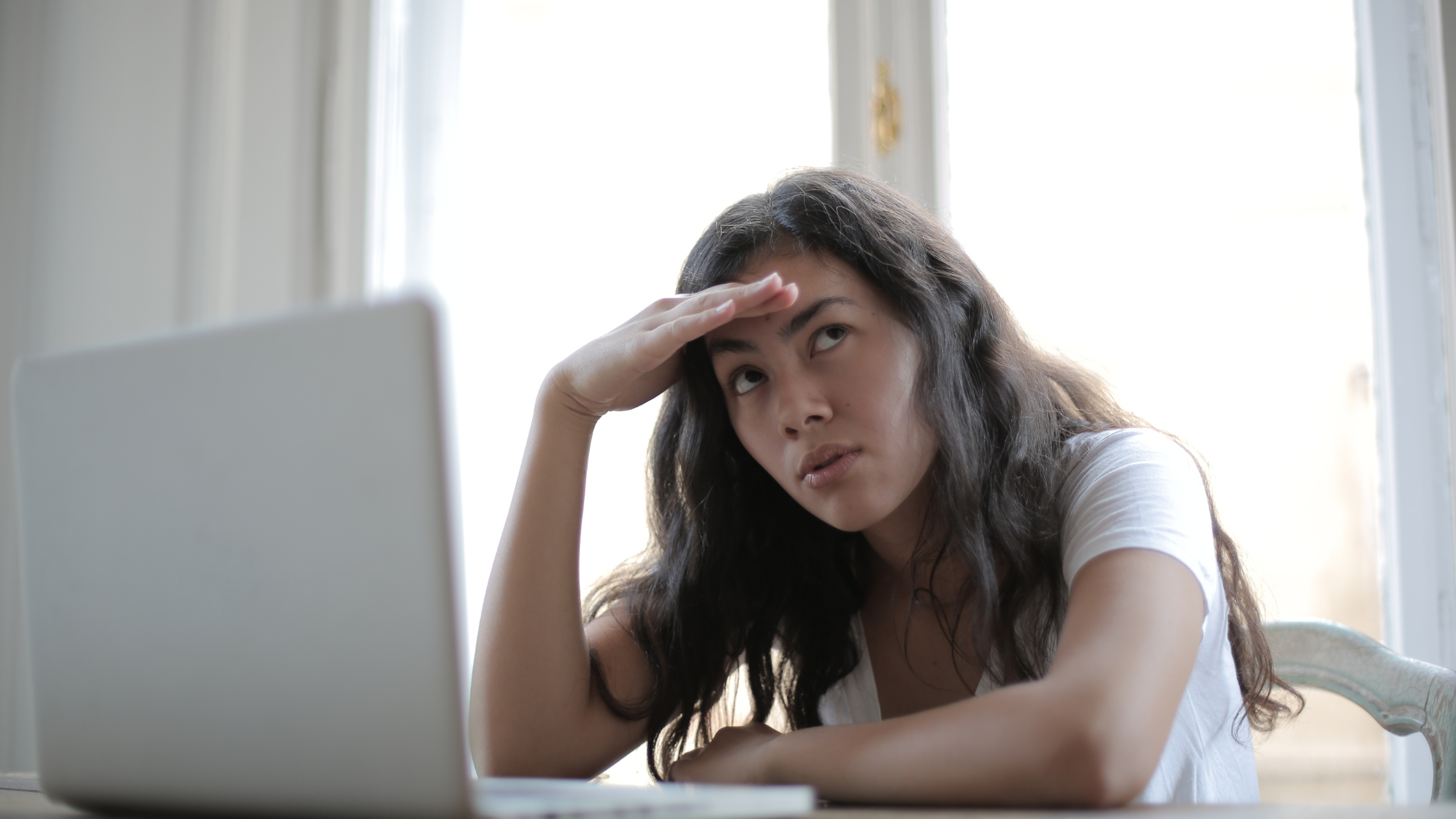 Woman holds head in hand, sitting at her laptop, confused in front of a large window