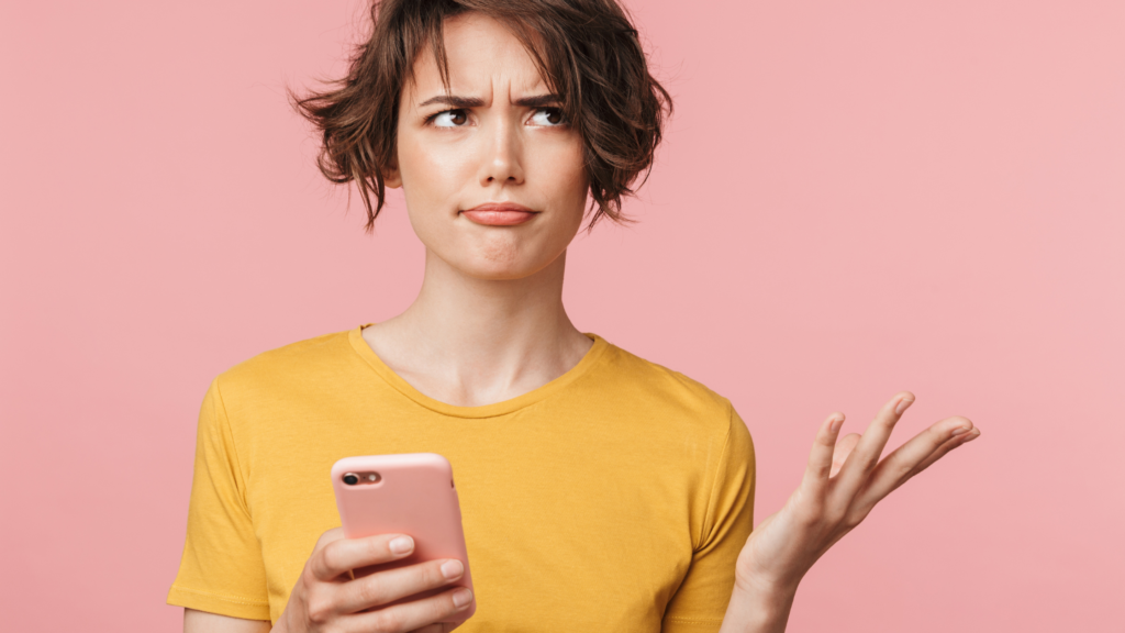 White woman with short brown hair wearing a yellow shirt, holds her pink smart phone and shrugs in confusion