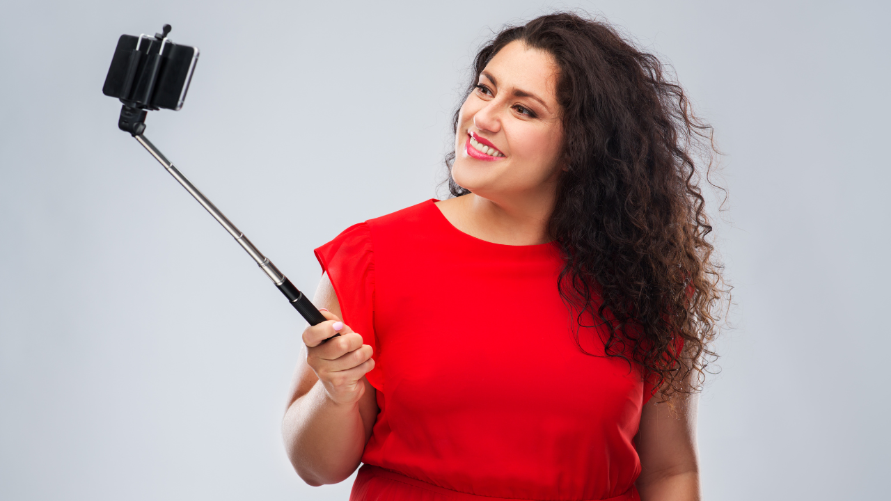 Woman with long, curly hair and wearing a red dress, holds a selfie stick and smiles into her phone camera